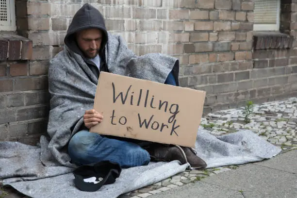 Homeless Man In Hood Sitting On Street Holding Cardboard With Text Willing To Work