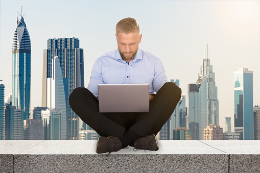 Young Businessman Sitting On The Rooftop Of A Skyscraper Using Laptop