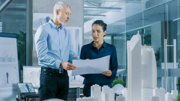 istock Two Professional Male and Female Architectural Engineers Work with Blueprints, Discussing Building Model Design for the Urban Planning Project.Clean Minimalistic Office, Concrete Walls Covered by Blueprints and Documents. 921019648