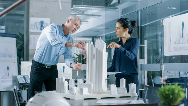 istock Two Professional Male and Female Architectural Engineers Work with Blueprints and on a Building Model Design for the Urban Planning Project.Clean Minimalistic Office, Concrete Walls Covered by Blueprints and Documents. 921019654