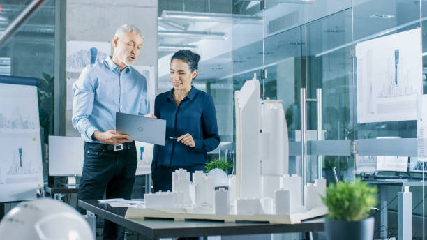 istock Male Senior Engineer Holds Laptop and has Discussion with Female Chief Engineer. They Work on a Building Model for a New District Urban Planning Project. 921019670