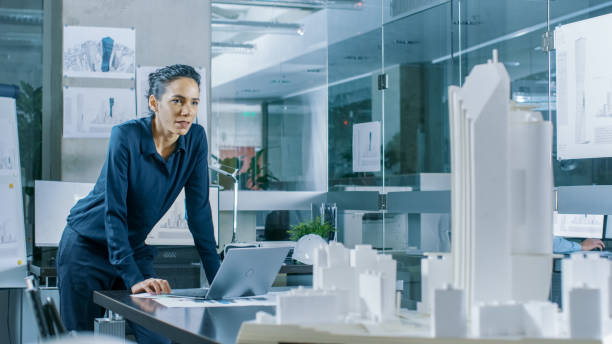 istock Female Architectural Designer Works on a Laptop,  Engineering New Building Model for the Urban Planning Project. Clean Minimalistic Office, Concrete Walls Covered by Blueprints and Documents. 921019680