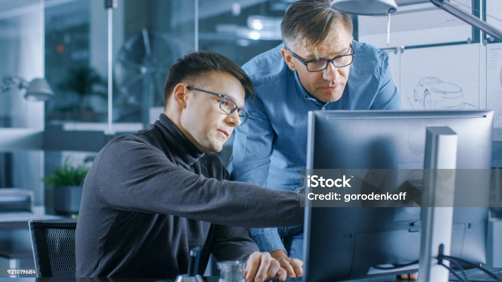 Experienced Senior Engineer Consults Young Designer about Project, They Have Discussion and Work on a Personal Computer. Engineer Stock Photo