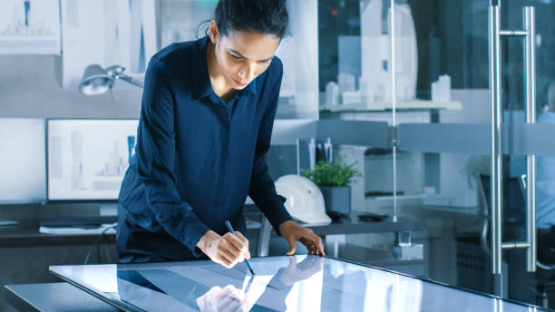 Talented Young Female Architectural Designer Draws Building Concept on a Graphics Tablet Display. Clean Minimalistic Office, Concrete Walls Covered by Blueprints.