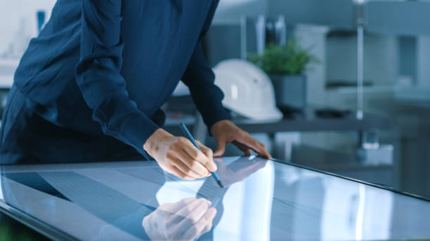istock Talented Young Female Architectural Designer Draws Building Concept on a Graphics Tablet Display Vertical Touchscreen Table. Clean Minimalistic Office, Concrete Walls Covered by Blueprints. 921019744