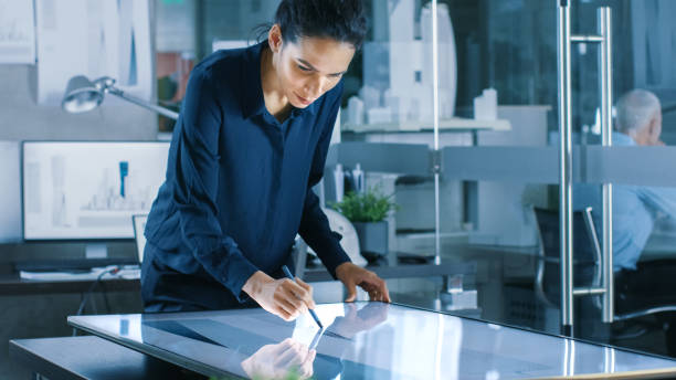 istock Talented Young Female Architectural Designer Draws Building Concept on a Graphics Tablet Display. Clean Minimalistic Office, Concrete Walls Covered by Blueprints. 921019750