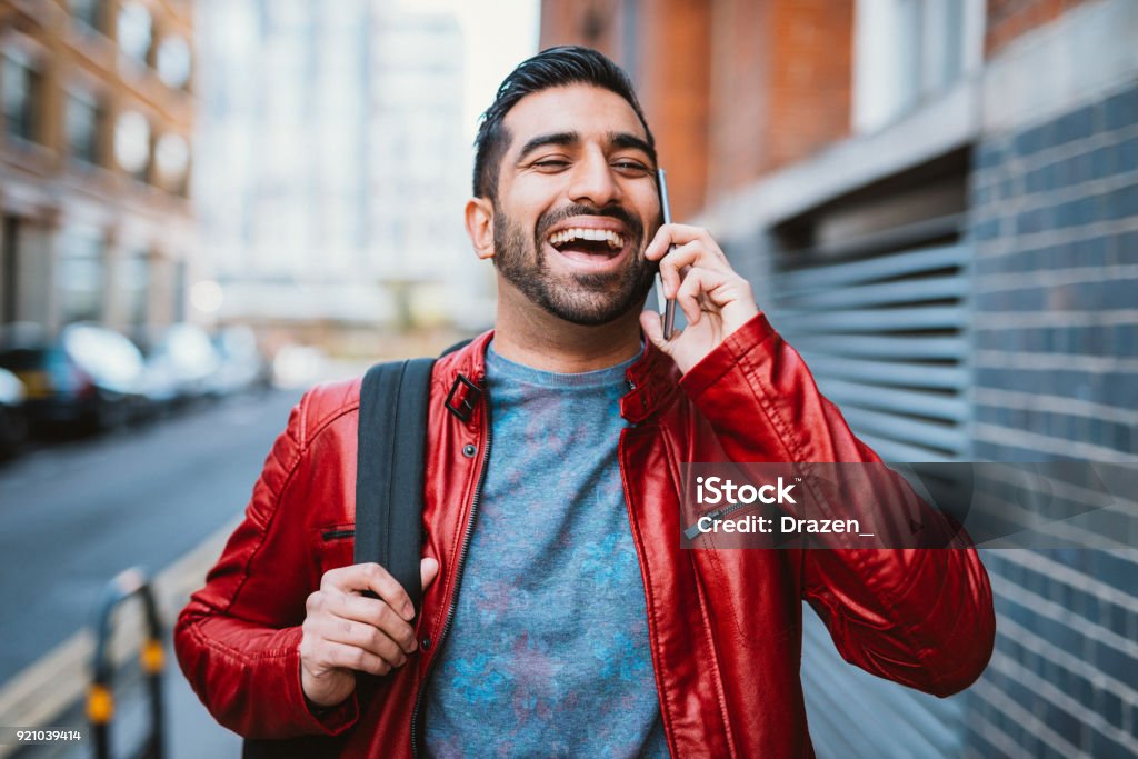 Man is using mobile phone to talk to friends Portrait of modern Indian guy in England, on autumn day in street style clothing. Cheerful 25 year old handsome man going to his workplace and enjoys building his career in creative business. Men Stock Photo