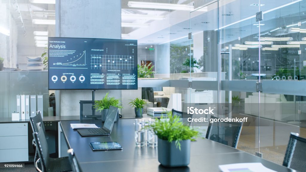 Modern Empty Meeting Room with Big Conference Table with Various Documents and Laptops on it, on the Wall Big TV Showing Company's Growth, Statistics and Pie Charts. Office Stock Photo