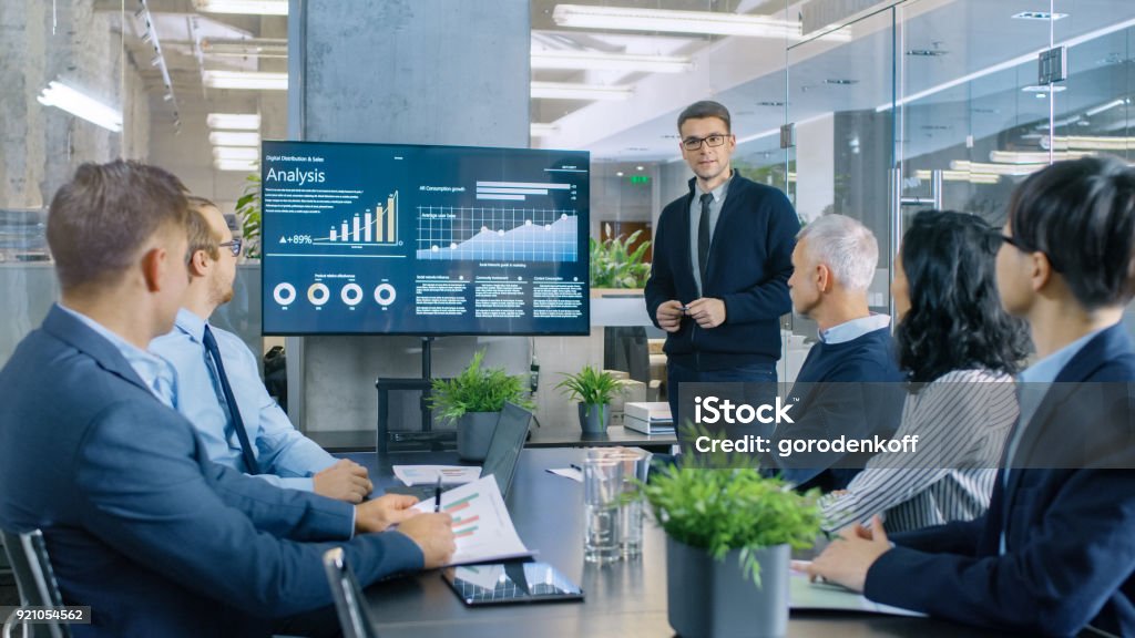 Young Stock Trader Shows to the Executive Managers Cryptocurrency and Trade Market Correlation Pointing at the Wall TV. Presentation - Speech Stock Photo