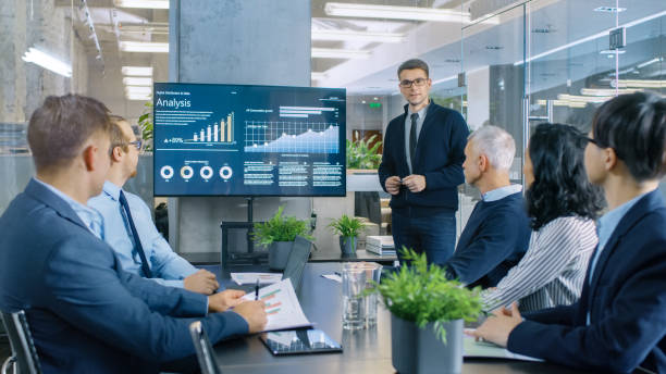 Young Stock Trader Shows to the Executive Managers Cryptocurrency and Trade Market Correlation Pointing at the Wall TV. Young Stock Trader Shows to the Executive Managers Cryptocurrency and Trade Market Correlation Pointing at the Wall TV. team meeting stock pictures, royalty-free photos & images