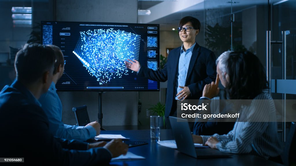 In the Conference Room Chief Engineer Presents to a Board of Scientists New Revolutionary Approach for Developing Artificial Intelligence and Neural Networks. Wall TV Shows Their Achievements. Meeting Stock Photo