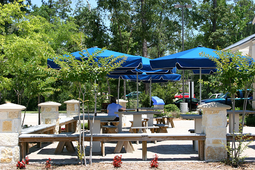Lady in blue sits beneath the blue umbrellas at the outside seating of a restaurant.  The area is surrounded by beautiful woodlands and shrubs.