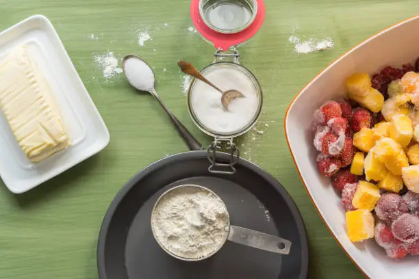 several types of baking utensils arranged on a green wooden surface with flour in a cub, sugar in a jar and frozen fruits in a bowl