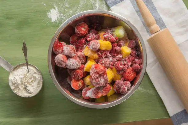 several types of baking utensils arranged on a green wooden surface with flour in a cub, sugar in a jar and frozen fruits in a bowl