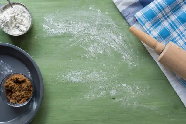 several types of baking utensils arranged on a green wooden surface with flour in a cub, sugar in a jar and frozen fruits in a bowl