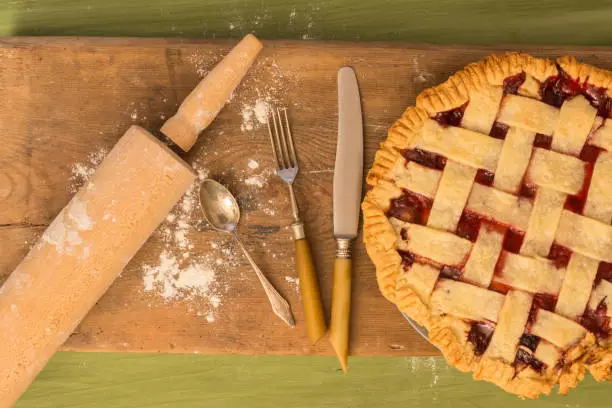 A pie on a wooden surface on a green table next to a rolling pin and some spilled flour