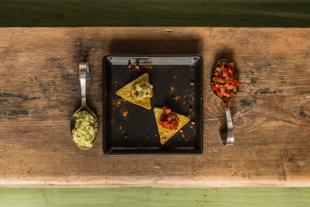 Nacho chips arranged on green colored wooden surface with lime, tomato, a spoon with paprika powder, a bowl of salsa and a bowl of guacamole