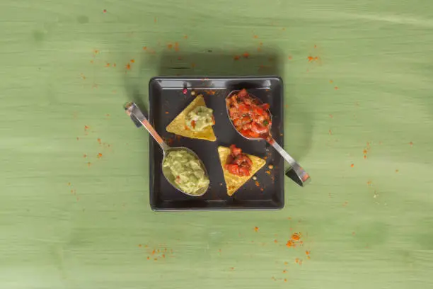 Nacho chips arranged on green colored wooden surface with lime, tomato, a spoon with paprika powder, a bowl of salsa and a bowl of guacamole