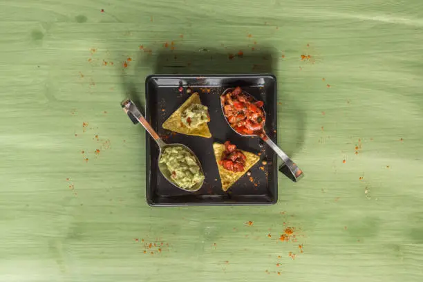 Nacho chips arranged on green colored wooden surface with lime, tomato, a spoon with paprika powder, a bowl of salsa and a bowl of guacamole