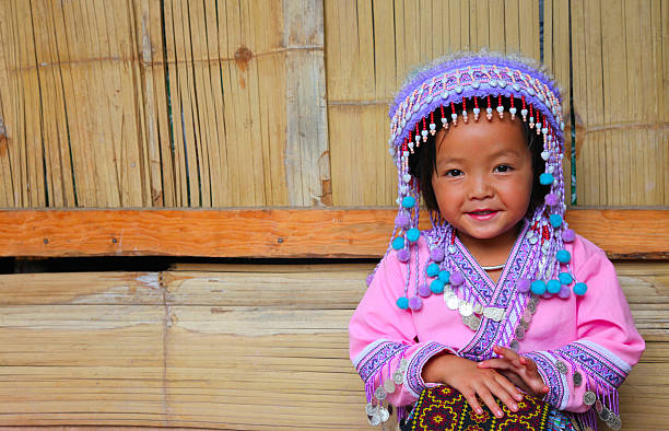 Rural town scene in north Thailand with traditioanlly dressed thai women. Women and some boys are on a simple road through town. One woman has also traditional hill tribe headwear and decosartions at clothings. Scene is in mountain town Pangkhum in Amphoe Samoeng in eastern Chiang Mai province. A man is passing by scene in foreground. At right side is a small old wooden building. In background is open mountain range landscape woth woodlands. A car is driving in left area in background