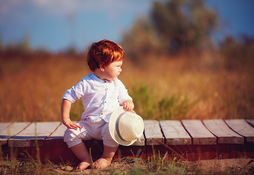 funny redhead toddler baby boy sitting on wooden path at summer field