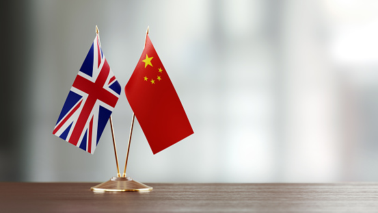 British and Chinese flag pair on desk over defocused background. Horizontal composition with copy space and selective focus.