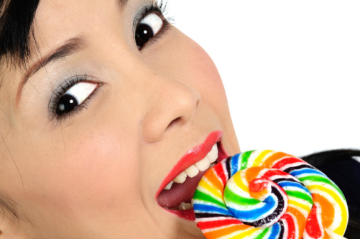 Side view of childish woman licking multicolor candy, wants to eat, looking at camera, showing tongue out, wearing casual style jacket. Indoor studio shot isolated on gray background.