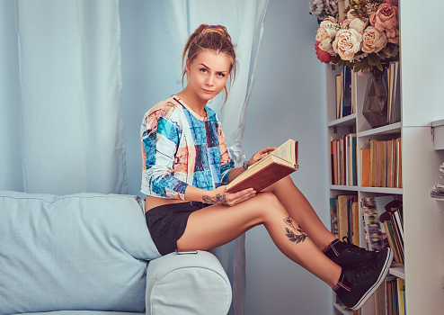 Young tattooed girl read a book sitting on a sofa at home.