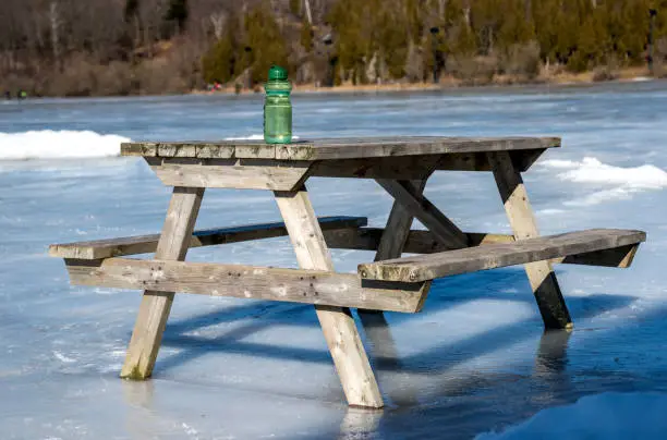 A wooden picnic table on a frozen lake. A green water bottle sits on top of the table. Sunny Day. Note horizon is not crooked, this is the shape of the land (bottle is straight).
