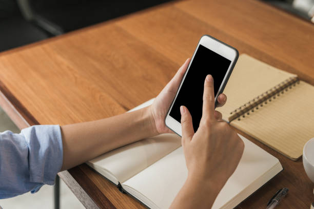 istock Young woman using smartphone mock up blank black screen while she working in the shop covered on a warmlight day. Young woman swipe her smartphone in a shop. indoor working in a house concept. 921294390