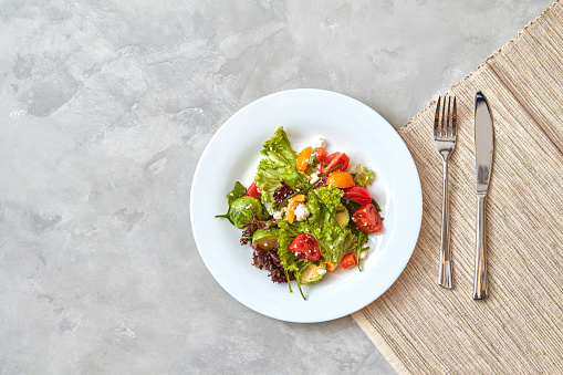 Portion Salad of fresh vegetables and cheese in a plate with a fork and knife on a gray background.step serving dishes, photo from Step by step recipe, Top view, flat lay.