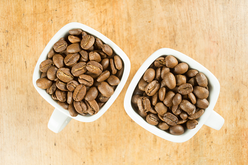 Light roast brown coffee beans in two white coffee cups on a wooden table top