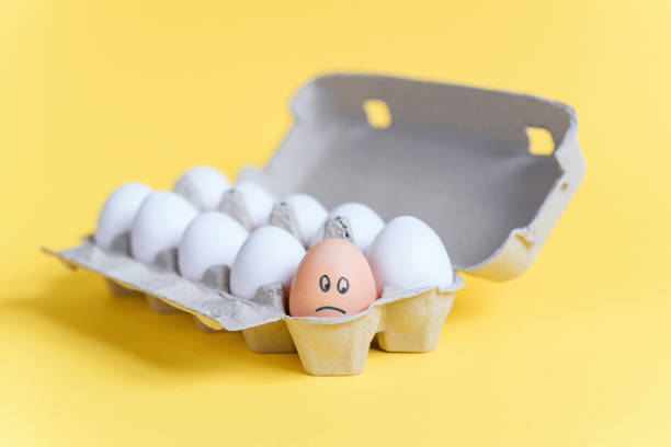 istock One orange egg with drawn sad face among white eggs in a cardboard tray on yellow background. 921426962