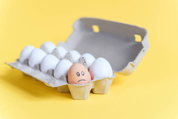 istock One orange egg with drawn sad face among white eggs in a cardboard tray on yellow background. 921426974