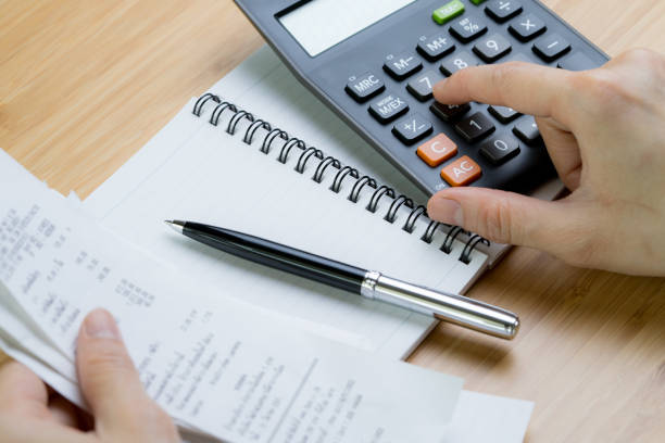 Cost calculation or bill payment concept, hand put finger on calculator and black pen on paper notepad with pile of bills in the left hand on wooden table stock photo