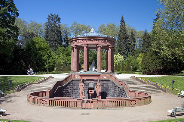 A tranquil garden landscape in Denmark showcasing a serene pond with a bronze statue, lush green grass, a weeping willow tree, and a peaceful white bench under a bright spring sky.