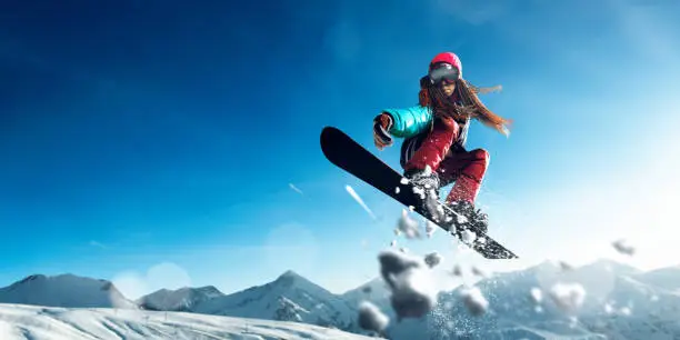 Female extreme snowboarder jumps and do a spectacular stunt in the air against a background of blue skies and snowy mountains. Freestyle snowboarding.