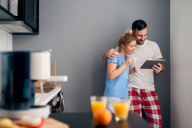 satisfecho sonriente hombre atractivo mostrando internet contenido a su bella novia curiosa en una tableta mientras ella bebe un café y de pie en la cocina en pijama. - standing digital tablet couple love fotografías e imágenes de stock