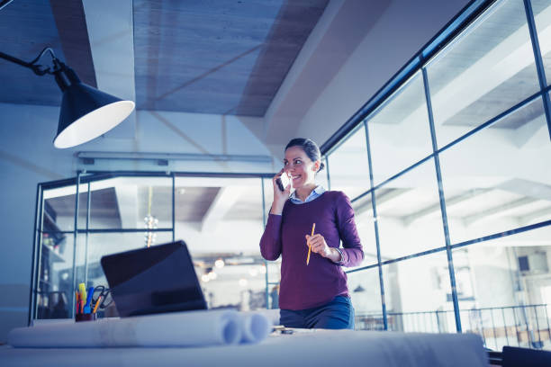 istock Young woman architect (designer), working on a project late at night, using mobile phone. Modern architectural studio 921587254