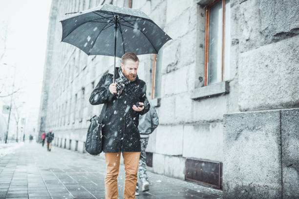 Ginger businessman walking in the snow, under the umbrella, using smart phone. Belgrade, Serbia, Europe