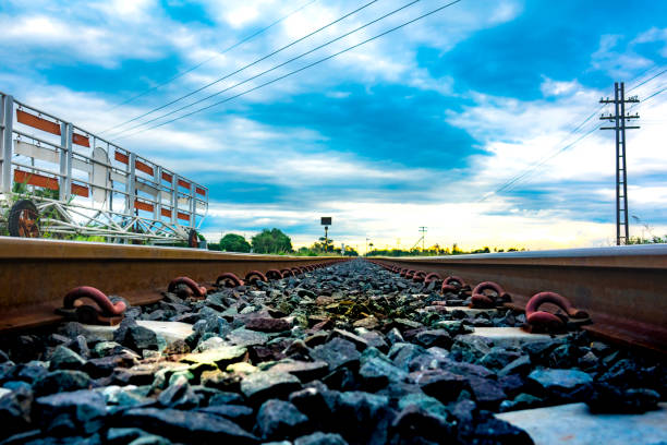 A view of railway tracks stretching into the distance, flanked by greenery and bushes in a rural area. The perspective emphasizes the long, straight path of the tracks as they lead towards distant hills under an overcast sky, capturing the essence of quiet countryside travel.