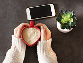 Young woman holding a heart shaped coffee cup, smartphone and plant