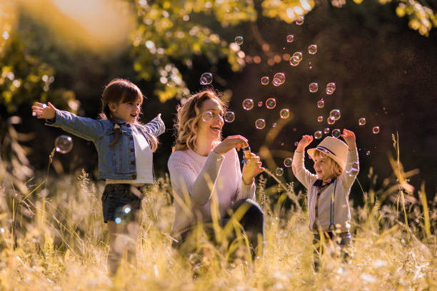 heureuse mère s’amuser avec ses enfants tout en soufflant des bulles dans l’herbe. - bubble wand outdoors little boys mother photos et images de collection