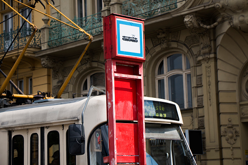 Close up of a tram stop sign with waiting tra and buildings in the background