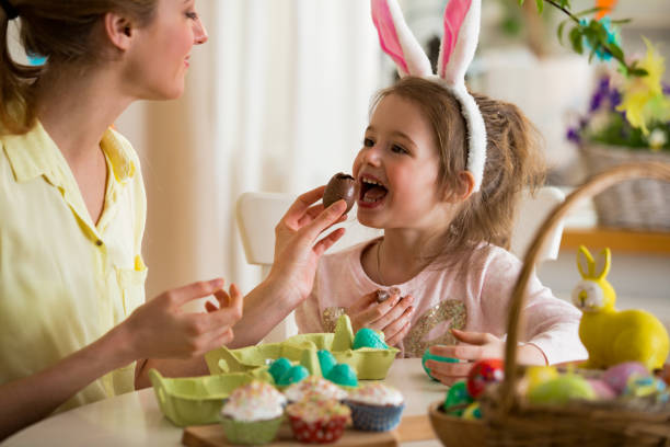 mutter und tochter feiert ostern schokoladeneier zu essen. glücklichen familienurlaub. niedliche kleine mädchen in hasenohren lachen, lächeln und spaß haben. - easter easter egg child chocolate stock-fotos und bilder