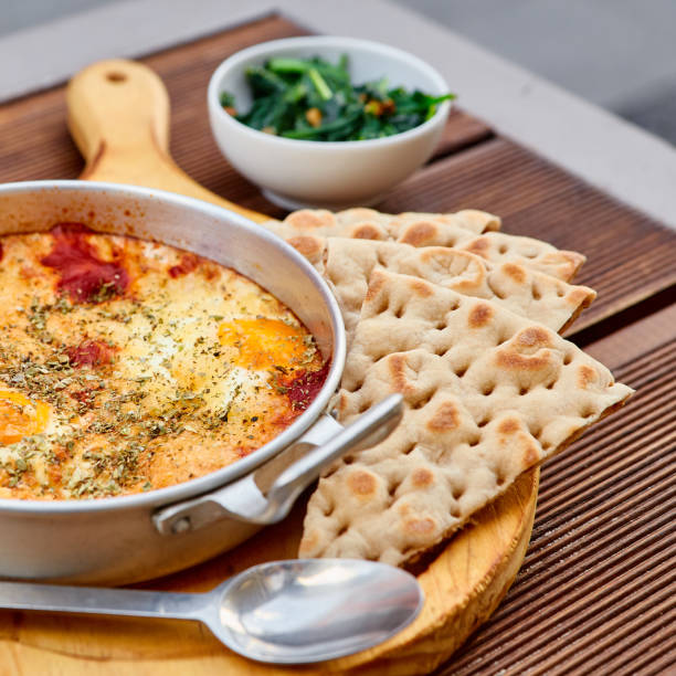 Scrambled eggs with tomatoes, cheese and spices in an aluminum frying pan with handles with homemade thin lavash and juicy greens on a wooden brown table. The photo was taken under natural light.