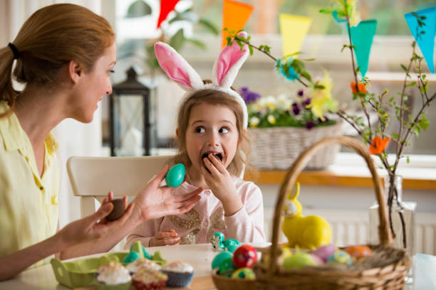 Mother and daughter celebrating Easter, eating chocolate eggs. Happy family holiday. Cute little girl in bunny ears laughing, smiling and having fun. Mother and daughter celebrating Easter, eating chocolate eggs. Happy family holiday. Cute little girl in bunny ears laughing, smiling and having fun. egg food photos stock pictures, royalty-free photos & images