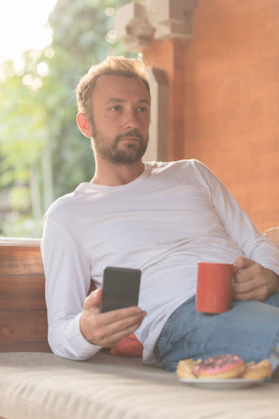 istock Man holding cellphone, coffe mug with donuts on the sofa. 921674160