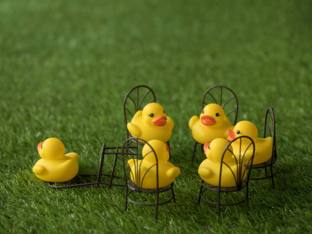 istock Group of five yellow rubber ducks sitting on chairs facing each other with one chair empty, knocked over and one duck running away, scene set on artificial grass. 921680888