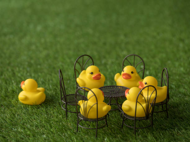 istock Group of five yellow rubber ducks sitting on chairs around a table with one chair empty and one duck running away, scene set on artificial grass. 921680890